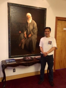 Michael Yen, Administrative Assistant to the Spiritual Assembly, stands next to a portrait of `Abdu'l-Bahá (son and successor of Bahá'u'lláh.